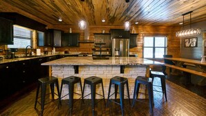 A kitchen with a large island, bar stools, and wooden ceiling. - Sleeps 24 * Luxe Pool Cabin Secluded w/Game Room (Broken Bow)