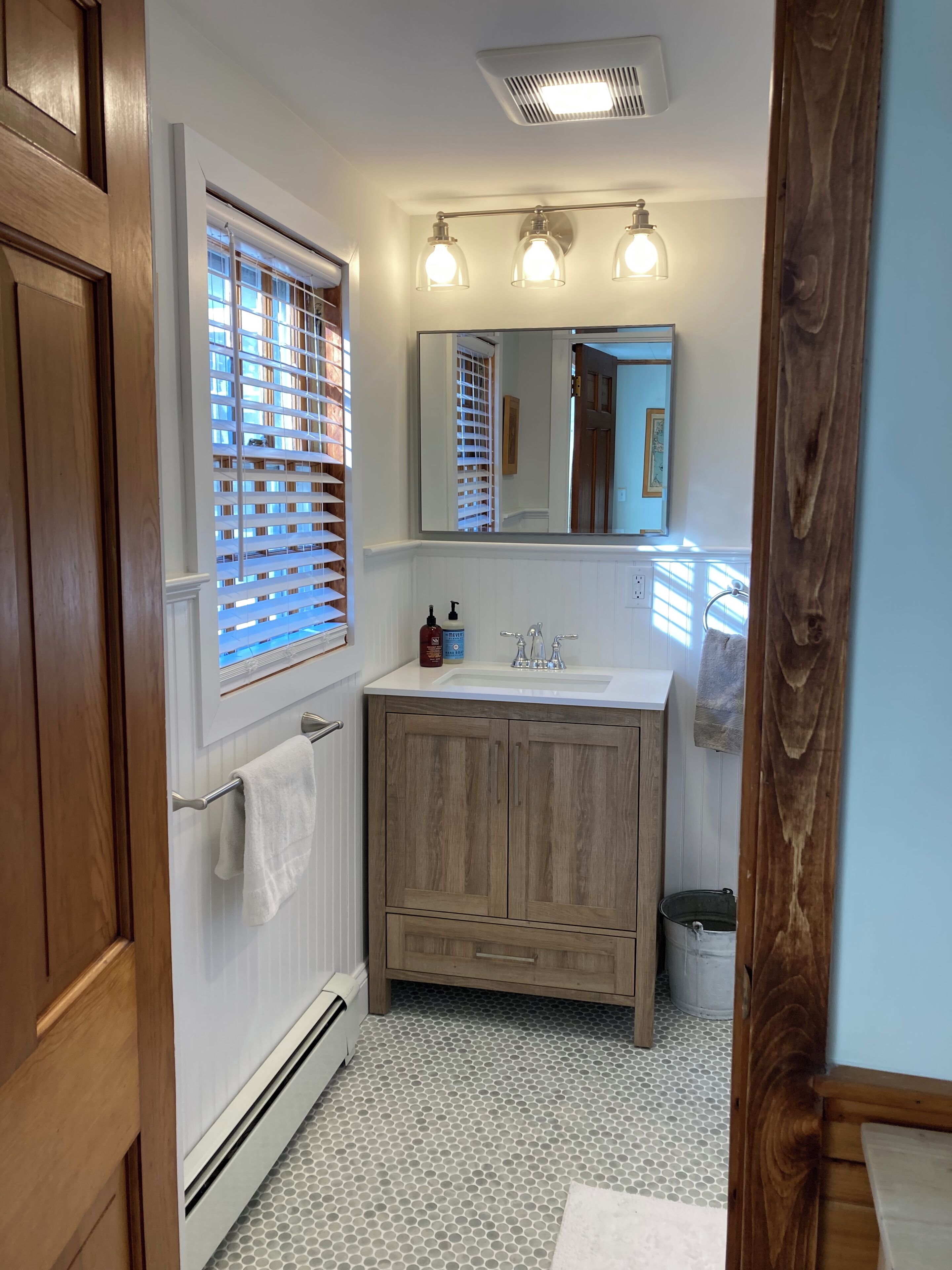 Un baño con tocador de madera, un espejo, una ventana con persianas y piso de baldosas.
