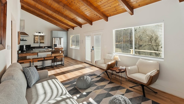 A spacious living area with a high wooden ceiling, a kitchen in the background, and a large window with blinds. - Peaceful Sunshine Chalet with Hot Tub (Big Bear Lake)