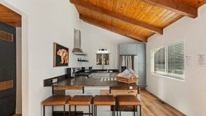 A modern kitchen with a wooden ceiling, a central island with bar stools, and a large window with blinds. - Peaceful Sunshine Chalet with Hot Tub (Big Bear Lake)