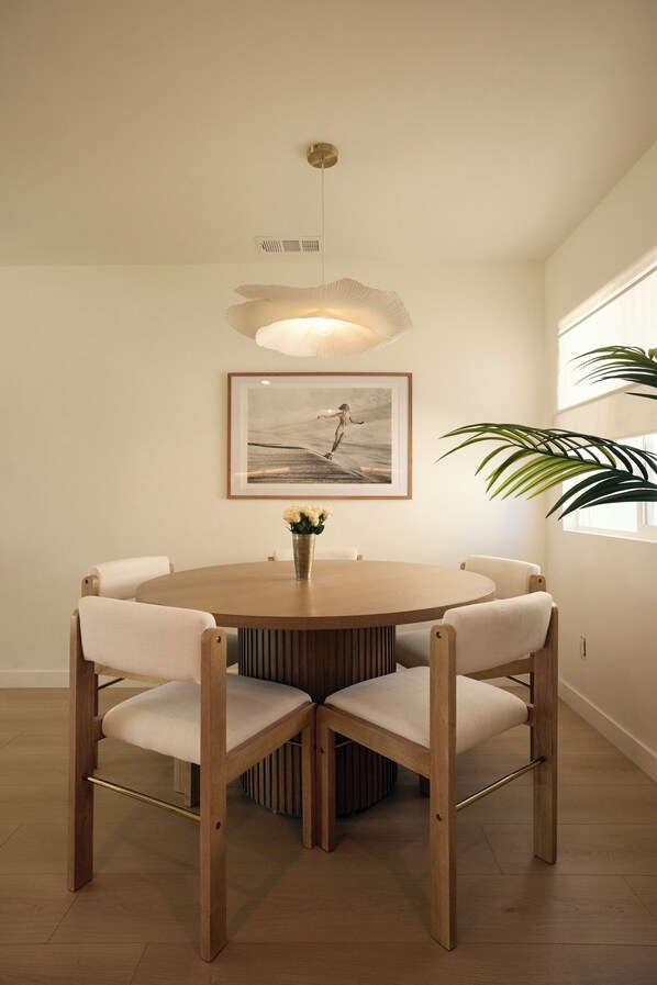 A round wooden dining table with four chairs, a framed picture on the wall, and a hanging light fixture.