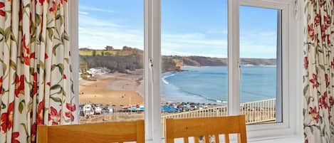 A room with a large window overlooking a beach and ocean, wooden chairs, and a table with a lamp.
