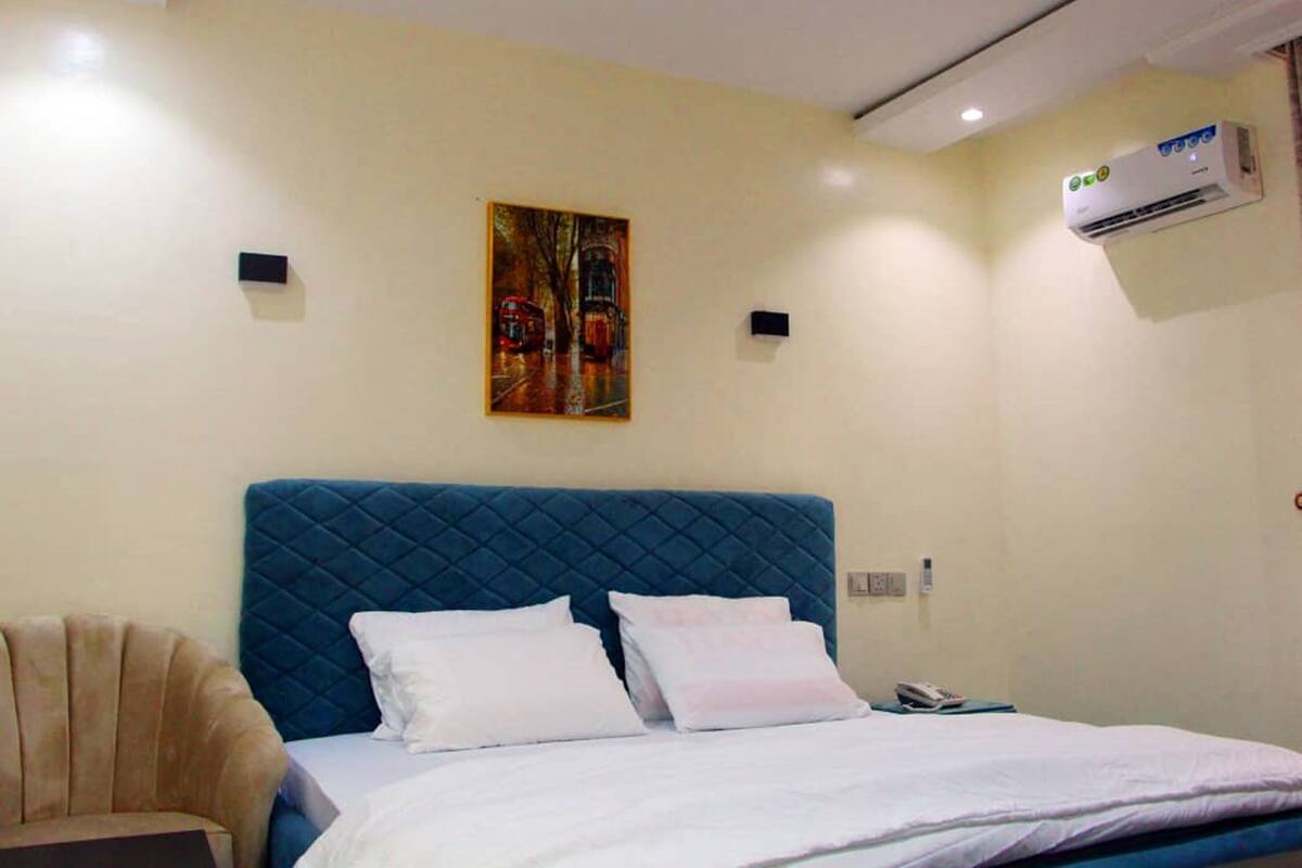 Una habitación de hotel con cabecera azul, cama con sábanas blancas, un cuadro en la pared, un equipo de aire acondicionado y una pequeña mesa con una silla.