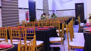 A banquet hall with rows of tables and chairs, a stage with floral arrangements, and a fire extinguisher on the wall. - S and B Suites (Lagos)