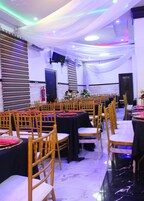 Uma sala de banquetes com fileiras de mesas e cadeiras, um palco com arranjos florais e um extintor de incêndio na parede.