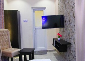 Quarto com televisão de tela plana na parede, poltrona acolchoada, mesinha lateral e planta em vaso.