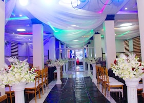 Espaço interior com um corredor central ladeado por colunas brancas e cadeiras, que leva a um palco.