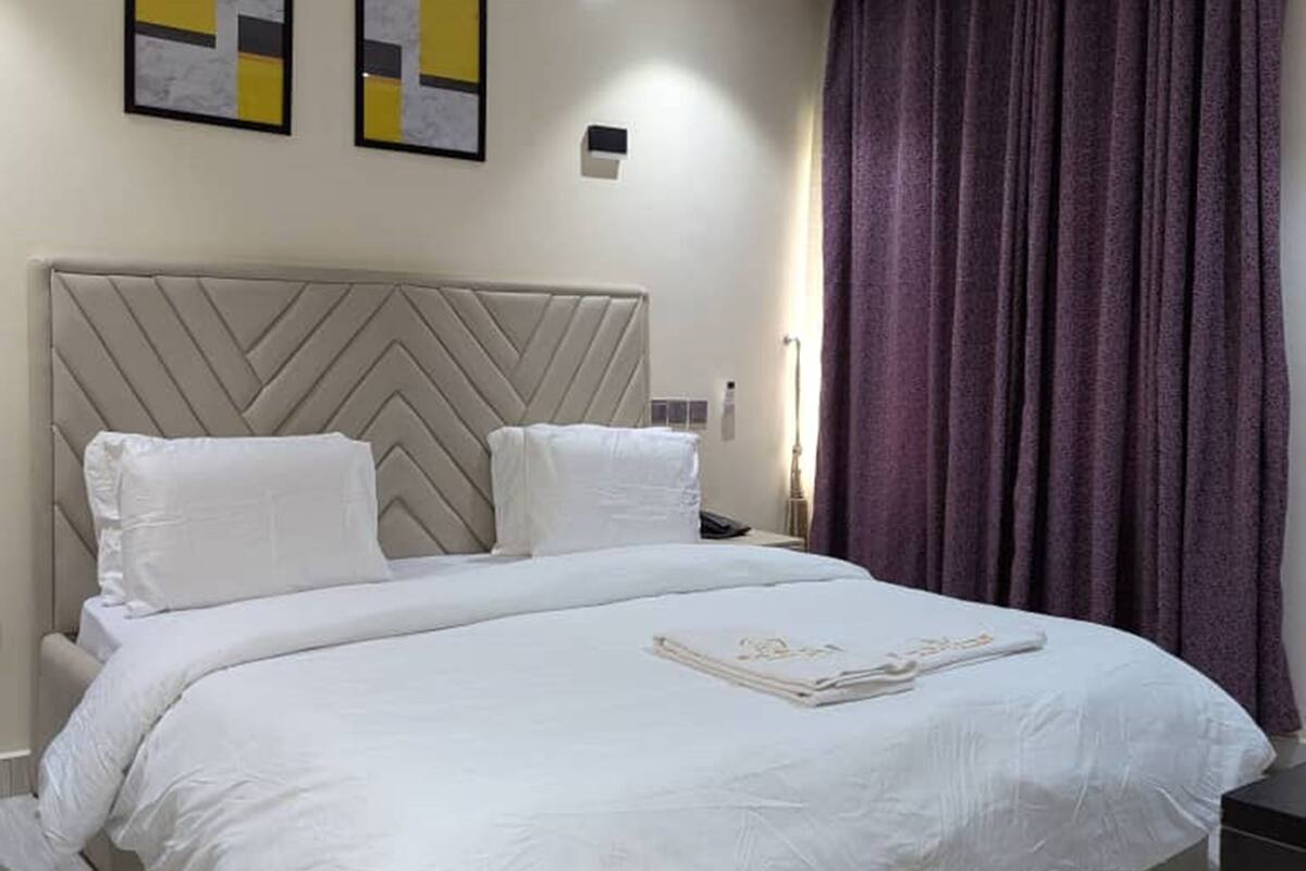Habitación de hotel moderna con una cama grande, cabecera con diseño geométrico, dos cuadros enmarcados en la pared y una cortina oscura en la ventana.