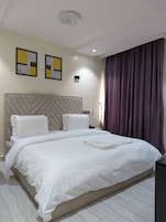 Quarto de hotel moderno com uma cama grande, cabeceira com padrão geométrico, duas obras de arte emolduradas na parede e cortina escura na janela.
