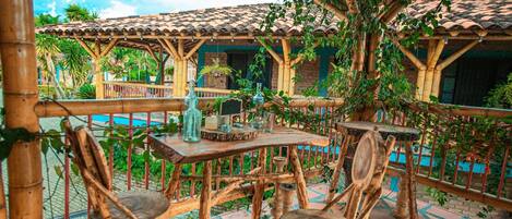 An outdoor patio with wooden tables and chairs, surrounded by bamboo structures and greenery.