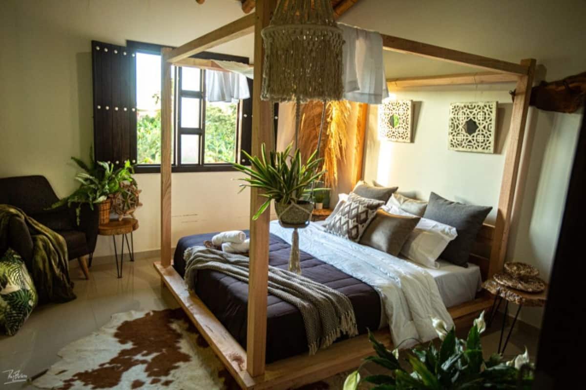 A four-poster bed with a wooden frame, a hanging macrame light fixture, and a view of the outdoors through a window with curtains.