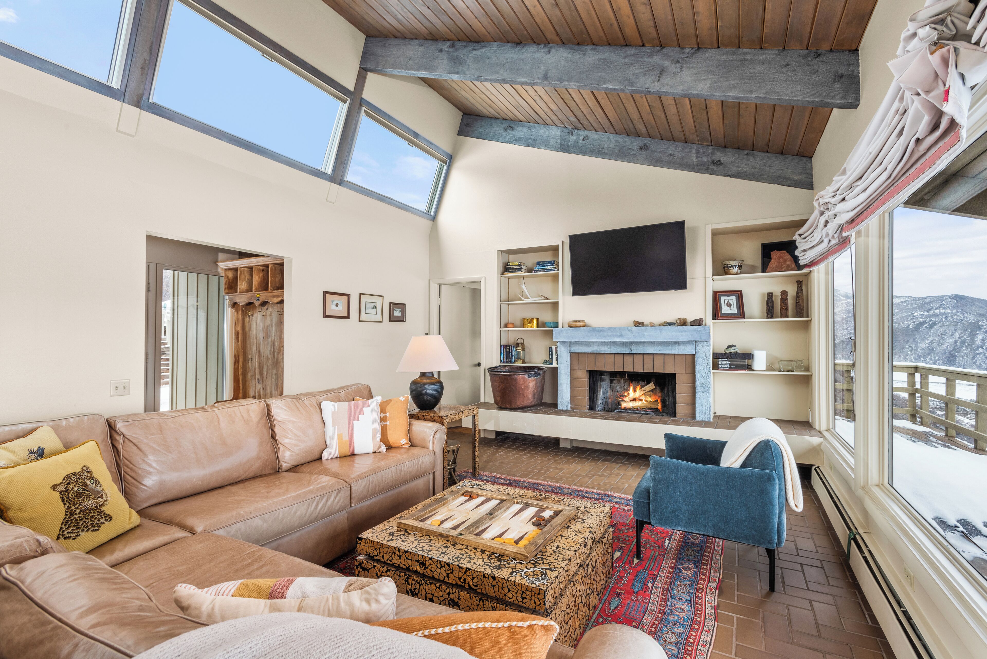 A spacious living room with a fireplace, leather sofa, and a large window overlooking a snowy landscape.