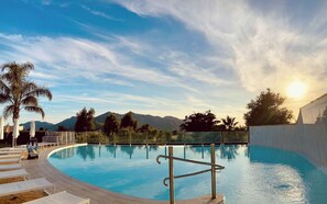 Une piscine offrant une vue dégagée sur les montagnes en arrière-plan, entourée de palmiers et dotée d’une terrasse avec des chaises longues.