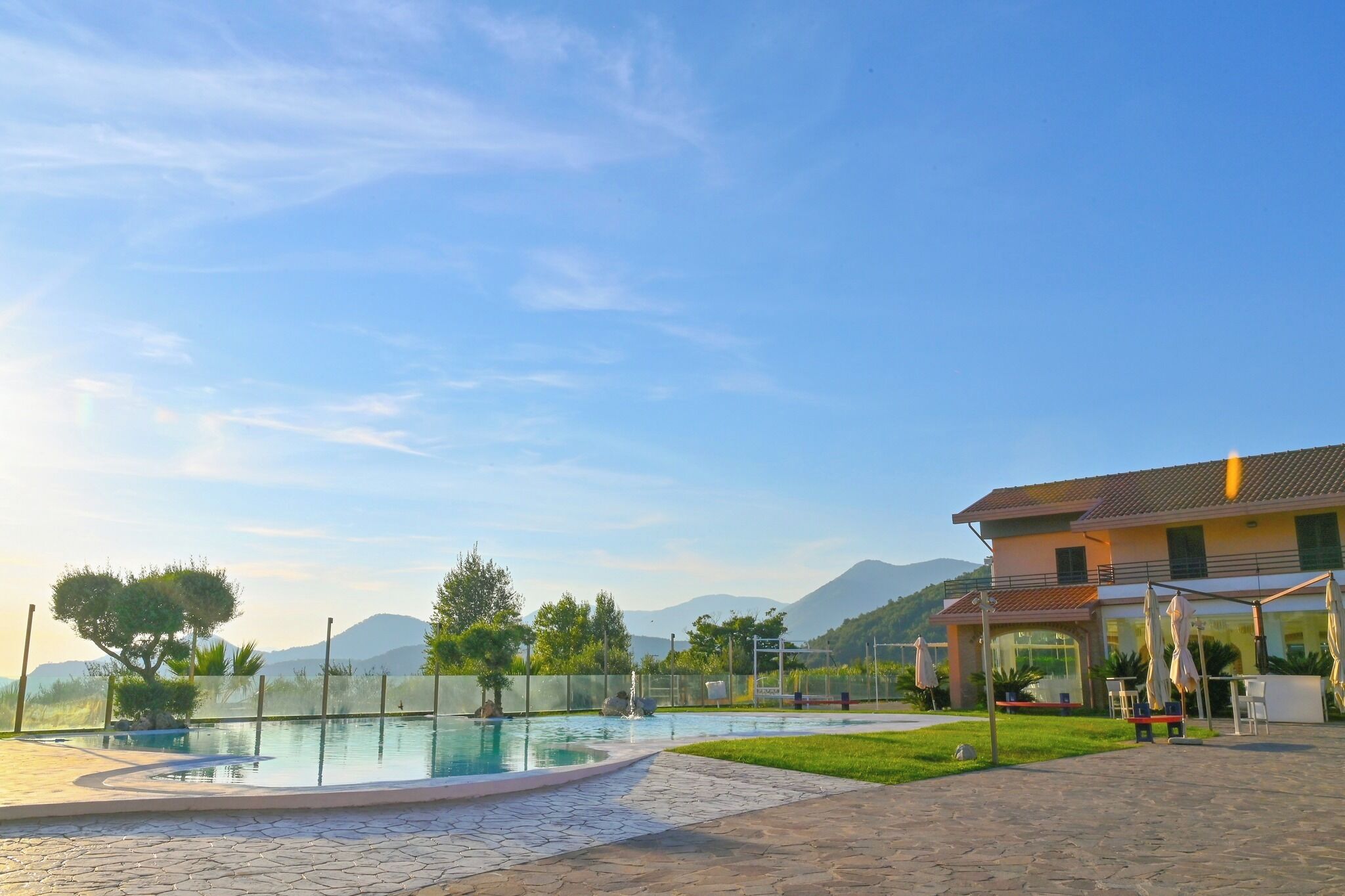 Une piscine avec une terrasse dallée, un bâtiment doté d’un espace extérieur aménagé, et un paysage montagneux en arrière-plan.