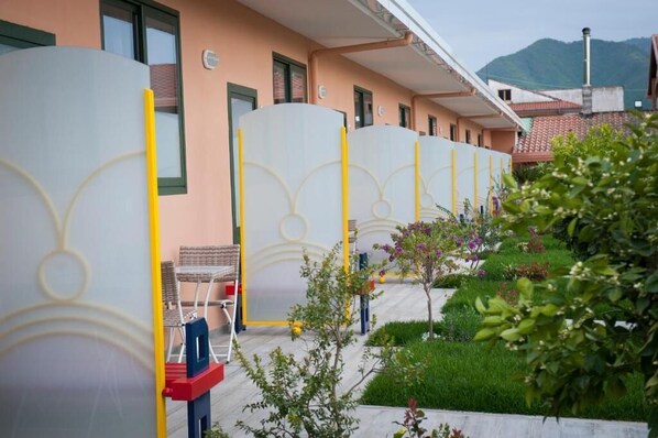 Une rangée de pièces séparées par de grandes cloisons en verre dépoli, un petit jardin agrémenté de plantes et de fleurs, et un bâtiment à la façade de couleur pêche.