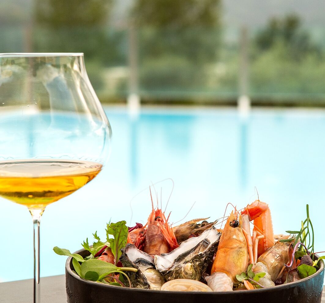 A glass of white wine next to a bowl of seafood including shrimp and oysters, with a pool in the background.