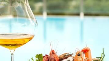 A glass of white wine next to a bowl of seafood including shrimp and oysters, with a pool in the background.