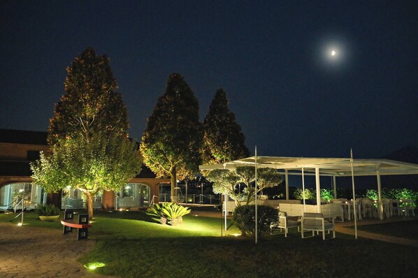 An outdoor area with a covered seating area, a bench, and a well-lit garden with trees.