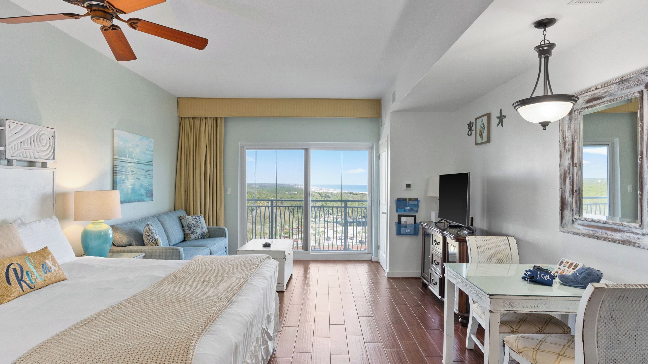 A hotel room with a bed, a sofa, a dining table, a ceiling fan, a TV, and a balcony with a view.