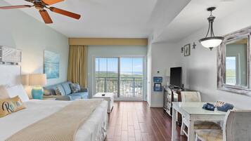 A hotel room with a bed, a sofa, a dining table, a ceiling fan, a TV, and a balcony with a view.