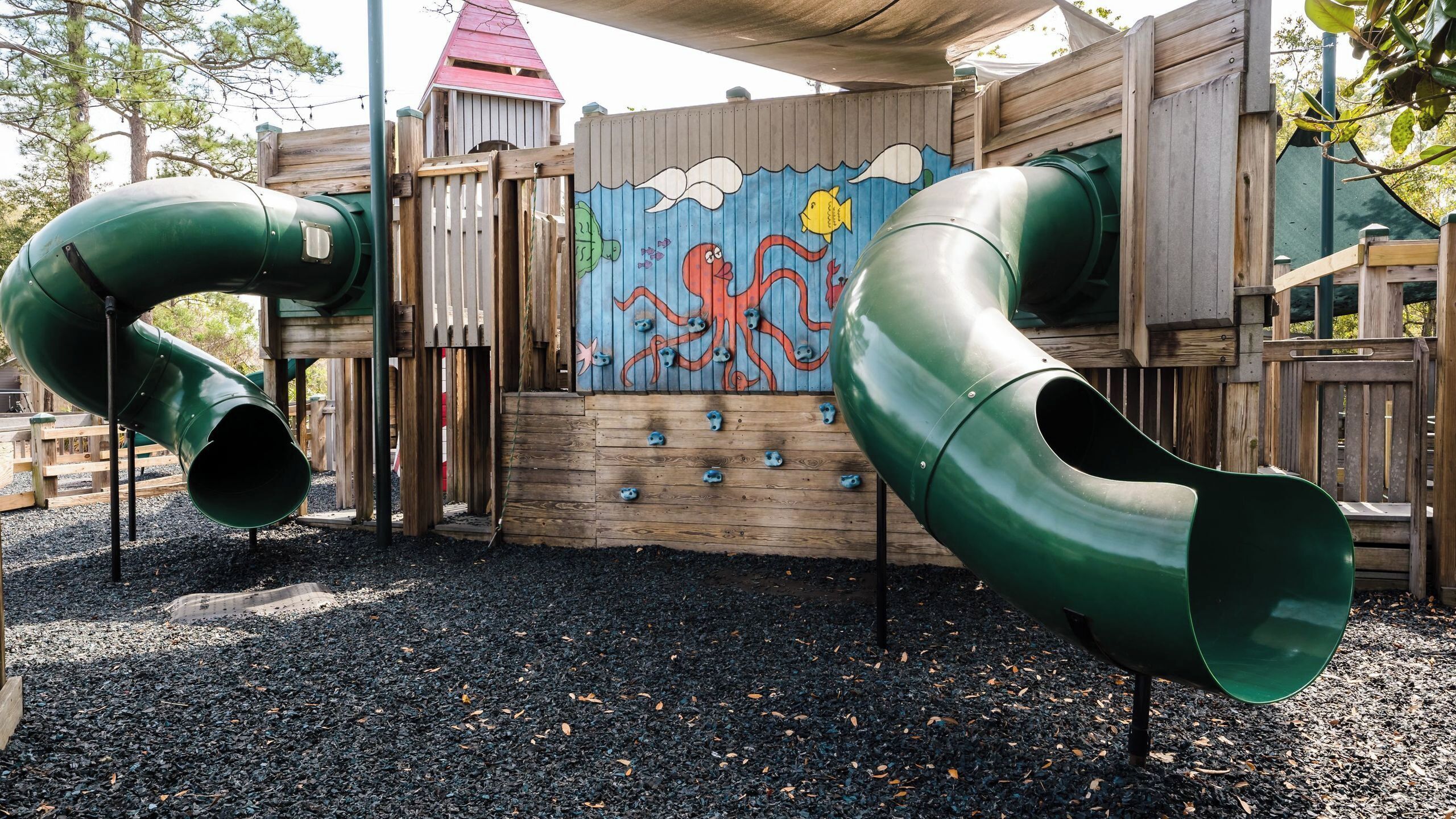 Un parque infantil con dos toboganes verdes y una pared de escalada de madera con un mural.