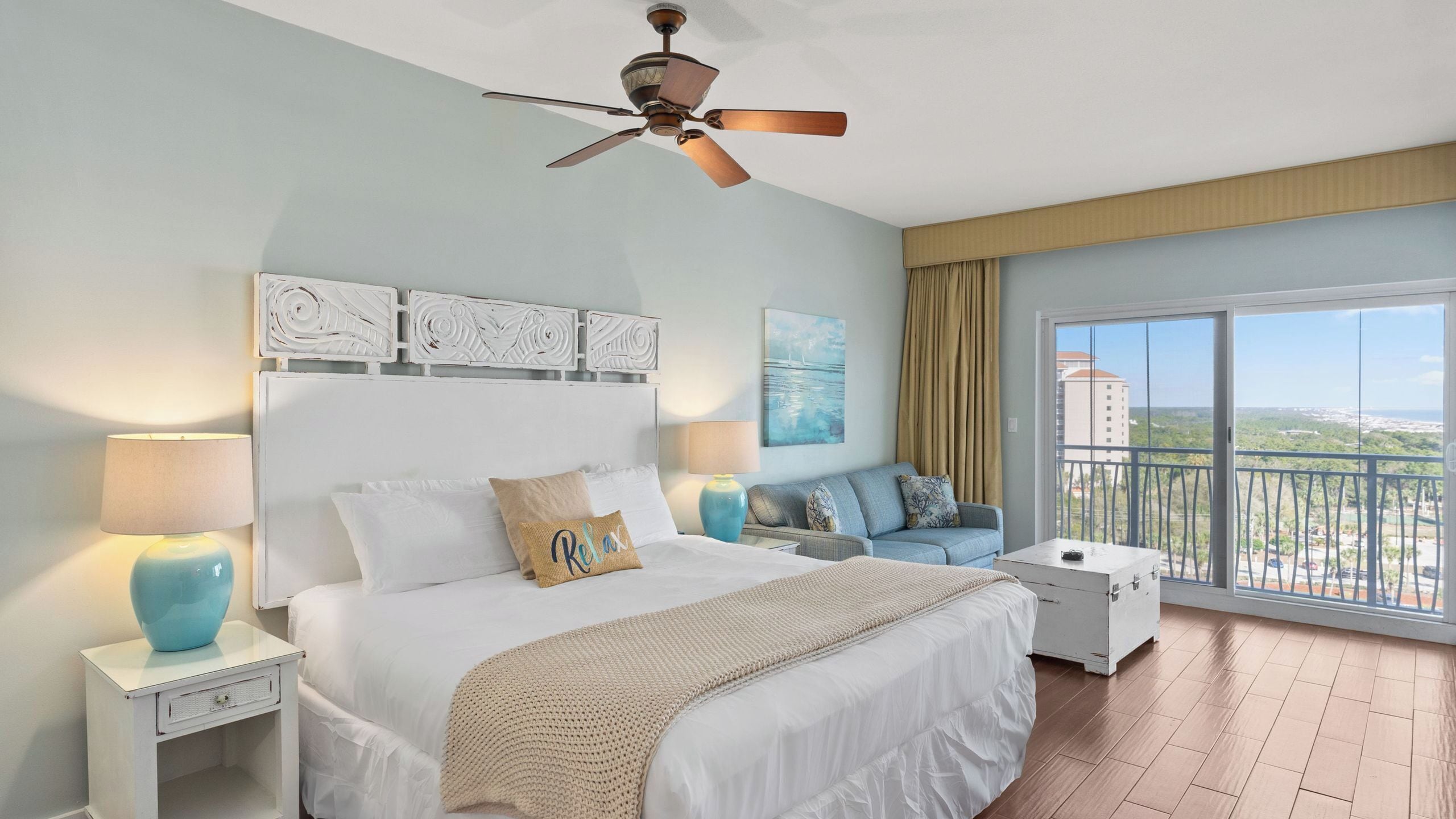 A bedroom with a bed, a sofa, a nightstand, a ceiling fan, and a large window with a view.