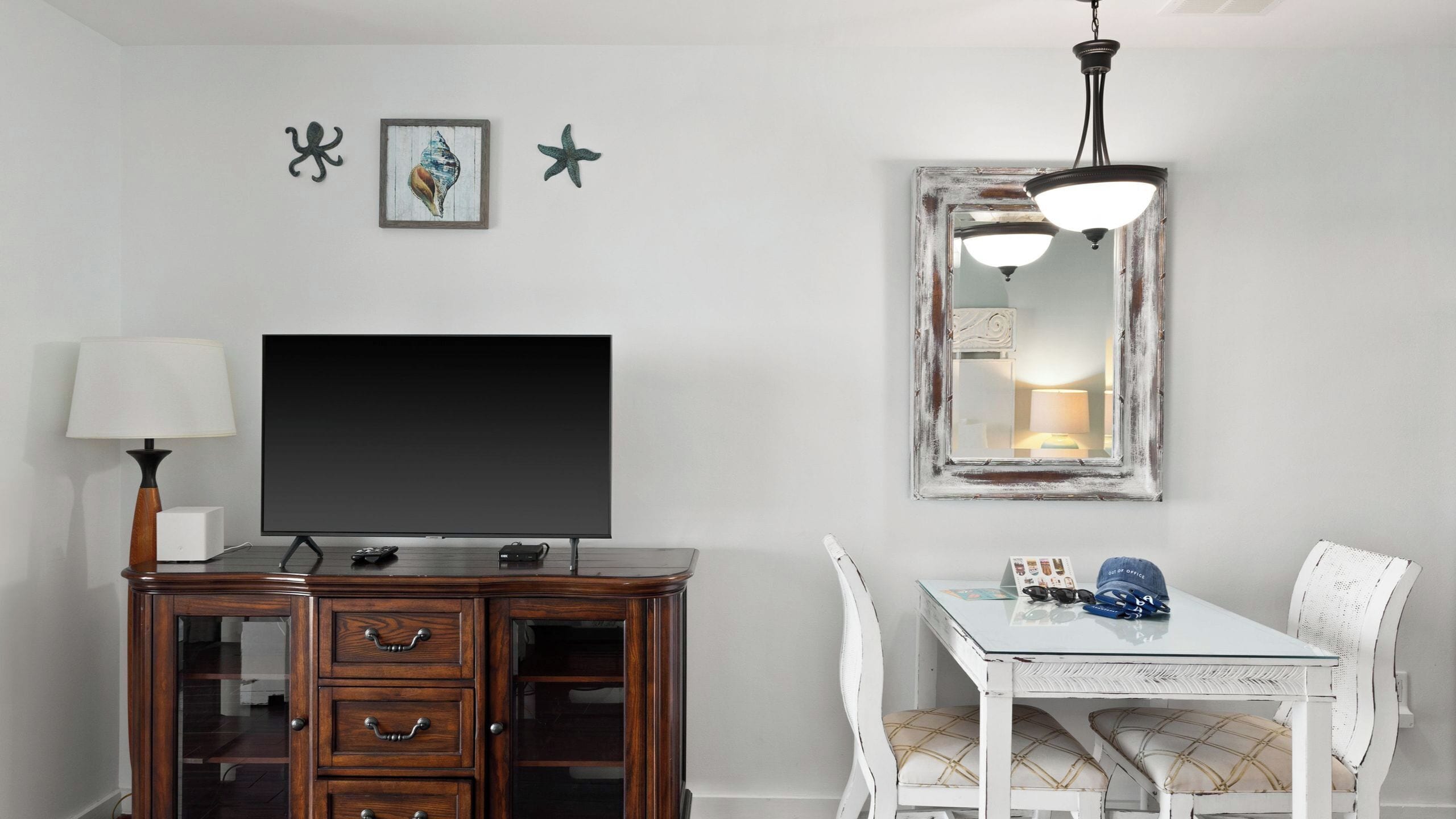 Une pièce comprenant une télévision, un meuble en bois, une table blanche, des chaises et un miroir avec un luminaire.