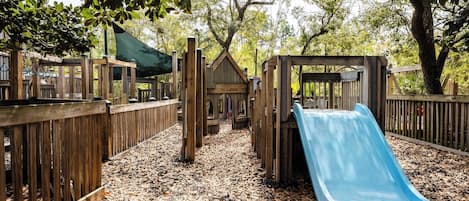 Un parque infantil con un tobogán azul, juegos de madera y una zona cubierta con asientos.