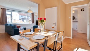 A dining area with a wooden table set for four, surrounded by chairs, with a vase of red flowers and wine glasses on the table. - Stylish 4 Bedroom Spacious House w/ Parking - Sleeps 10 - Families & Contractors (Broad Oak)