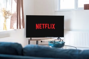A television with the Netflix logo on a stand in a room with a window, a radiator, and a vase with dried flowers.