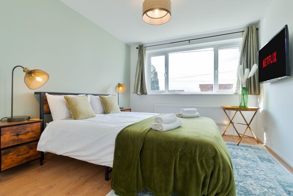 A neatly made bed with a green blanket, two bedside lamps, a TV on the wall, and a window with curtains.