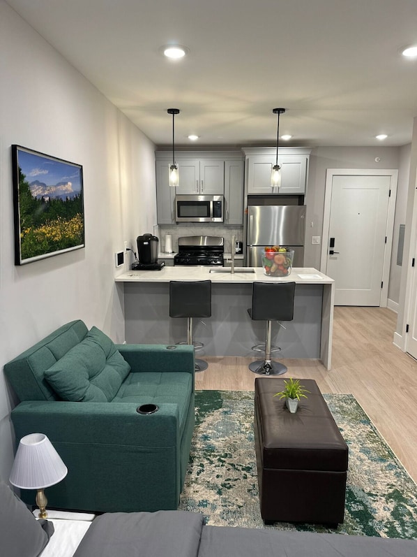 A modern living room with a green sofa, a coffee table, and a kitchen area with grey cabinets, a microwave, and a refrigerator. - Modern 1BR 1BA Apartment (Jersey City)