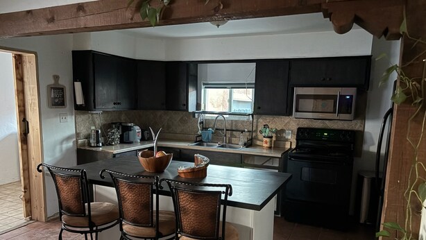 A kitchen with a central island, four bar stools, dark cabinets, a microwave, and a sink. - Cozy North Valley Charm (Albuquerque)