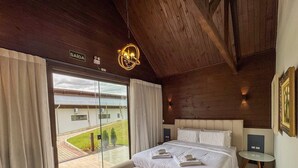 Un dormitorio con una cama grande, techo de madera, vista al exterior y una pequeña mesa con lámpara.