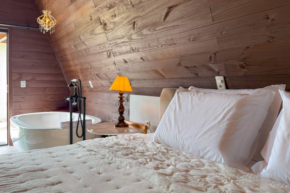 Una camera da letto con soffitto in legno, un letto con lenzuola bianche, un comodino con una lampada e una vasca da bagno.