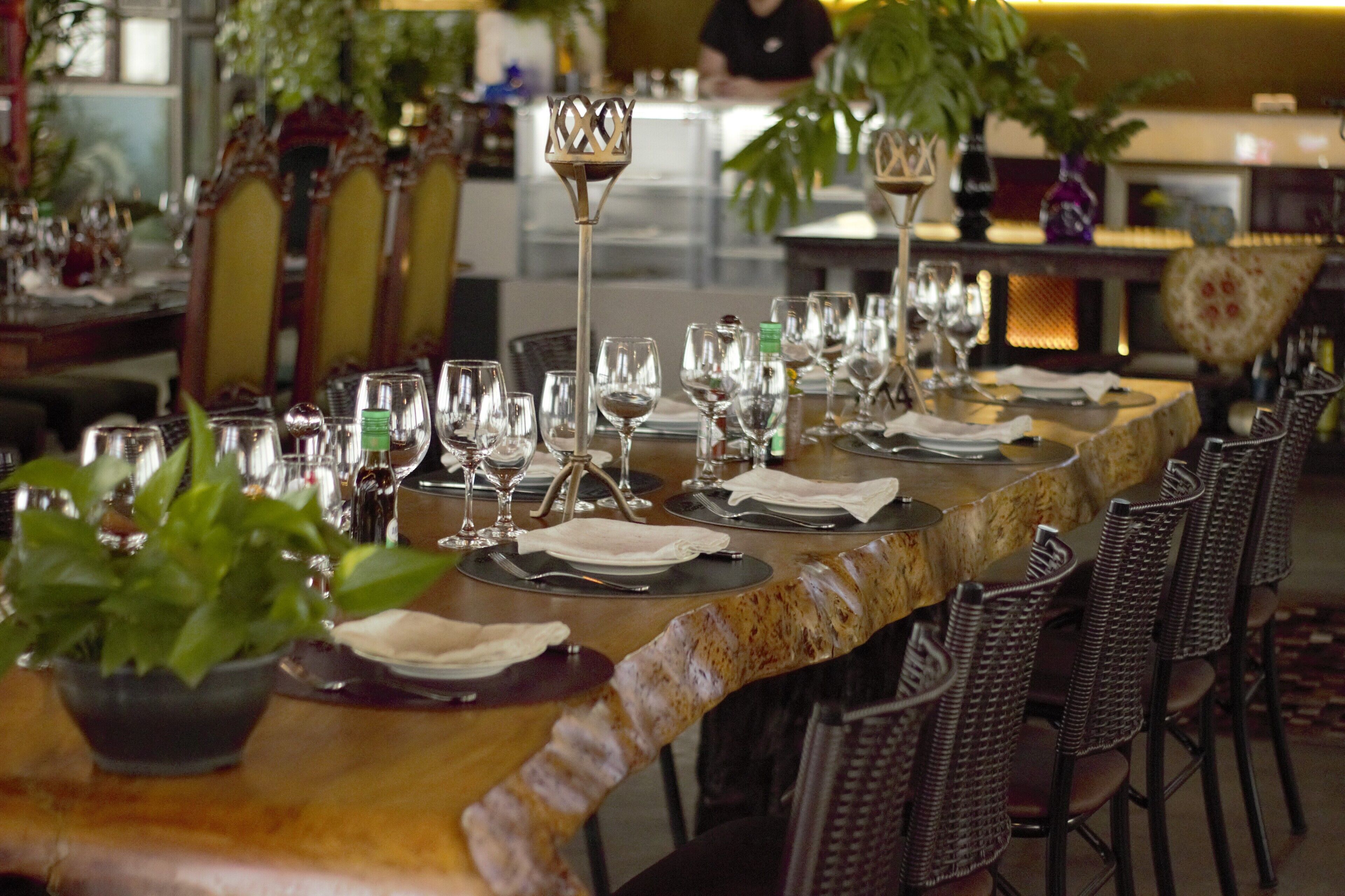 Une longue table à manger, dressée avec de nombreux verres à vin, des assiettes et des couverts, entourée de chaises en osier.