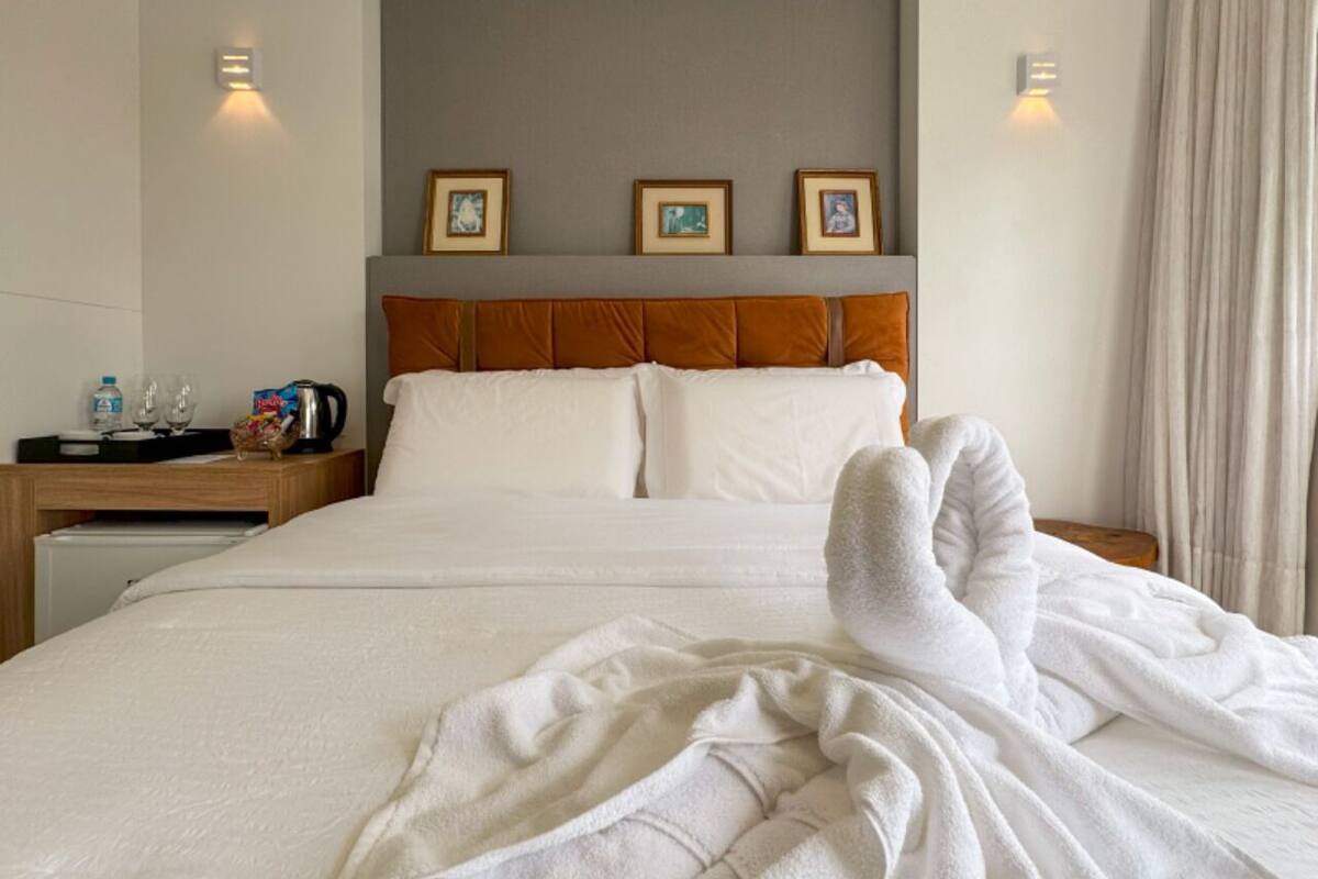 Un letto ben rifatto con un copriletto bianco e cuscini, un comodino in legno con bollitore e bicchieri, e quadri appesi al muro.
