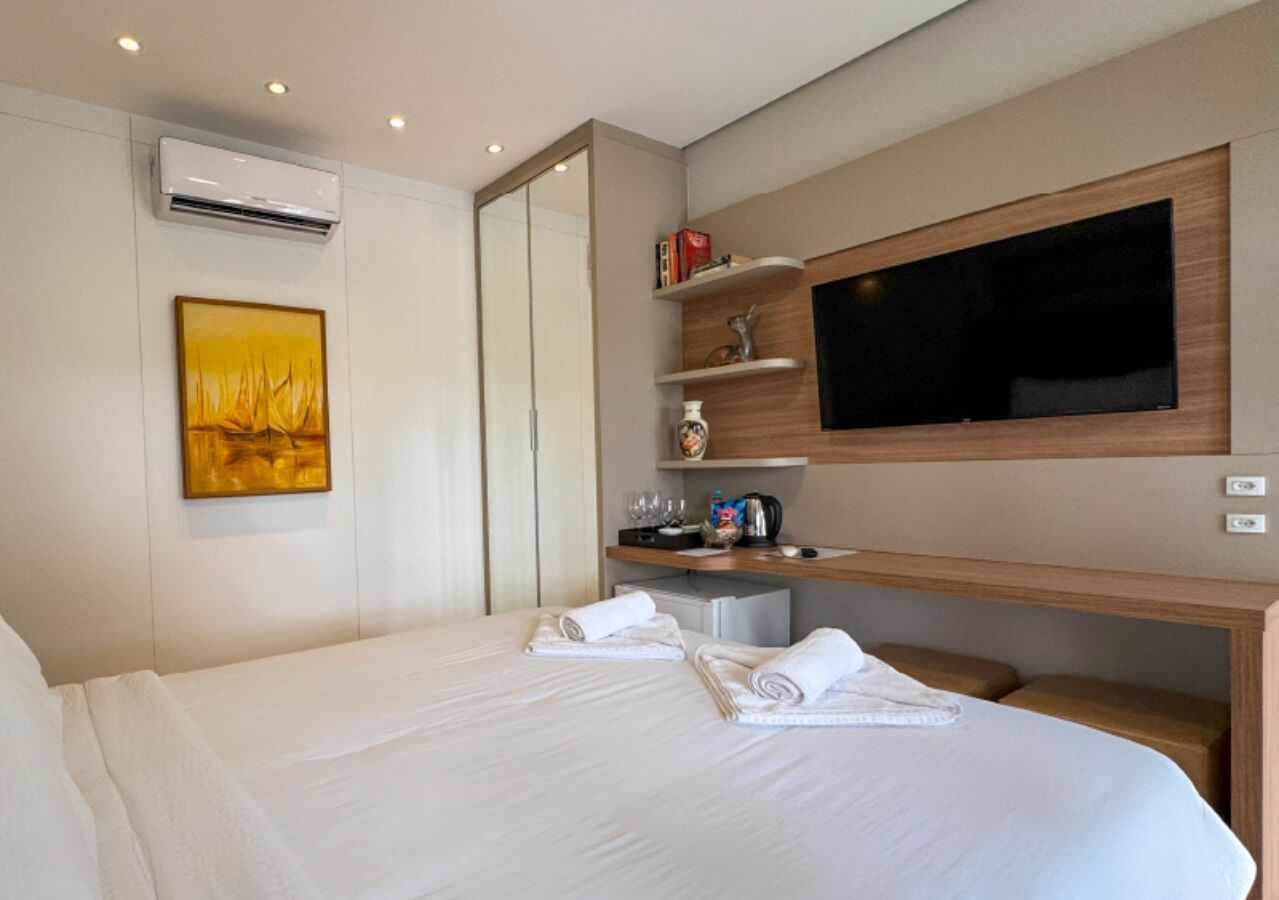 Quarto de hotel com uma cama grande, uma televisão de ecrã plano na parede, uma pequena secretária com uma chaleira e uma estante com objetos decorativos.