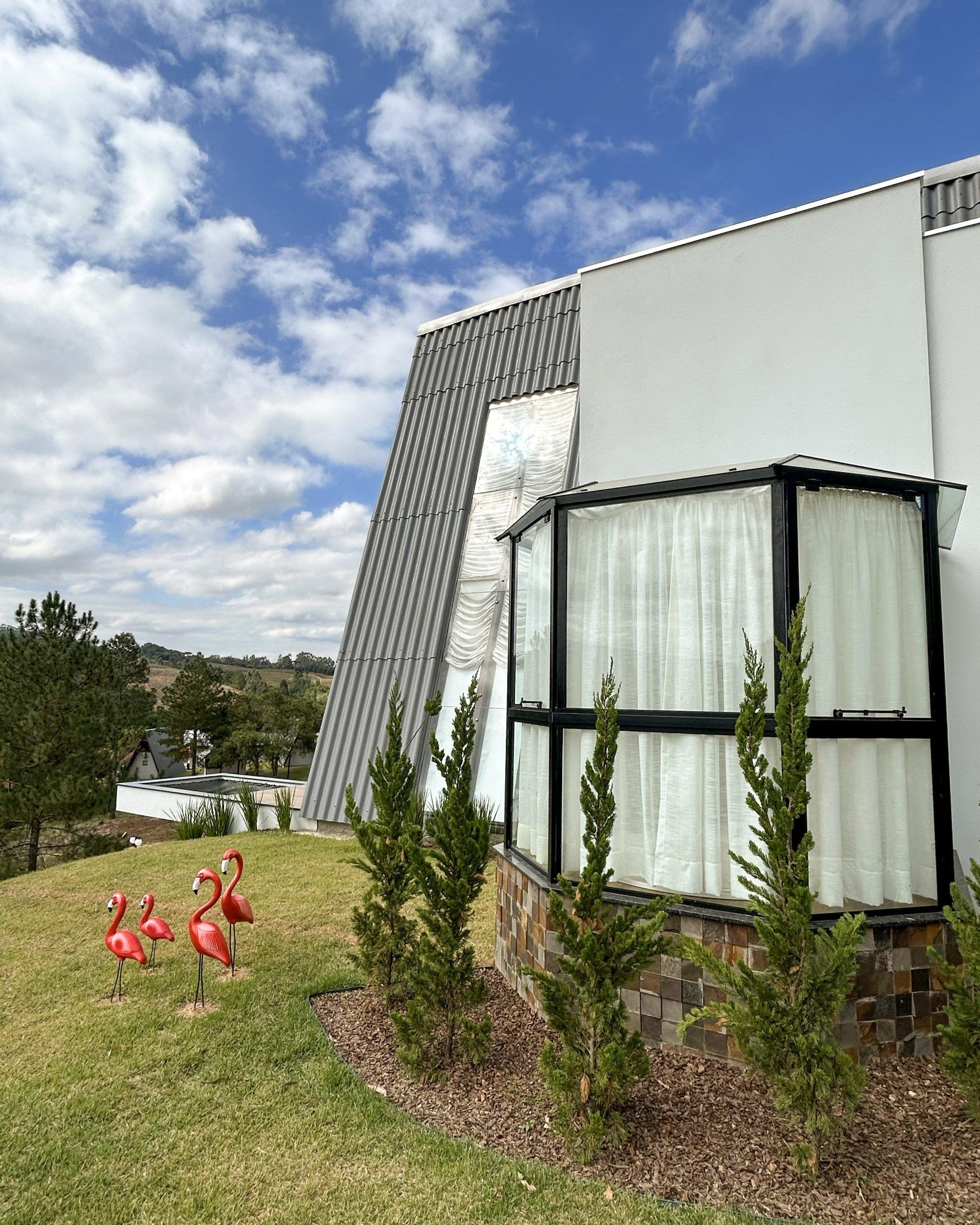 A modern house with a sloped roof, a large window with white curtains, and a small garden with flamingos and pine trees.