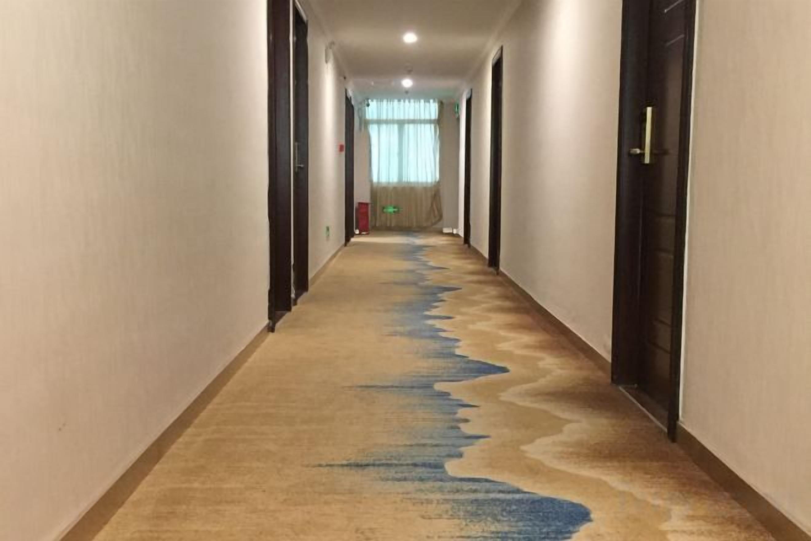 A hotel hallway with a carpeted floor, doors on both sides, and a window at the end.