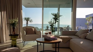 A modern living room with a sofa, armchair, and coffee table, featuring a view of the sea and mountains.
