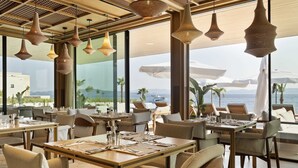 A restaurant with tables set for dining, large windows offering a view of the sea, and hanging pendant lights.
