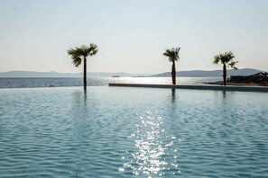 A pool with clear water reflecting sunlight, surrounded by palm trees and a distant view of the sea and mountains.