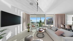 A modern living room with a large flat-screen TV, a sofa set, a dining area, and a view of the cityscape through large windows.