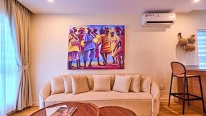 A living room with a beige sofa, two round wooden tables, a painting on the wall, an air conditioner, and a window with blinds. - Stone House Apartment  (Abuja)