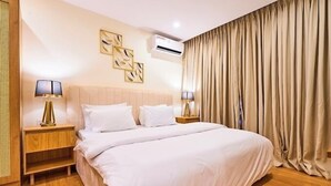 A hotel room with a large bed, two bedside tables with lamps, a wall-mounted air conditioner, and a large window with beige curtains. - Stone House Apartment  (Abuja)