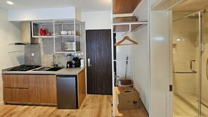 Een compacte keuken met een inbouwfornuis, een spoelbak en een compacte koelkast. Er is een houten kast met een hangrek en een klein houten kistje op de vloer.