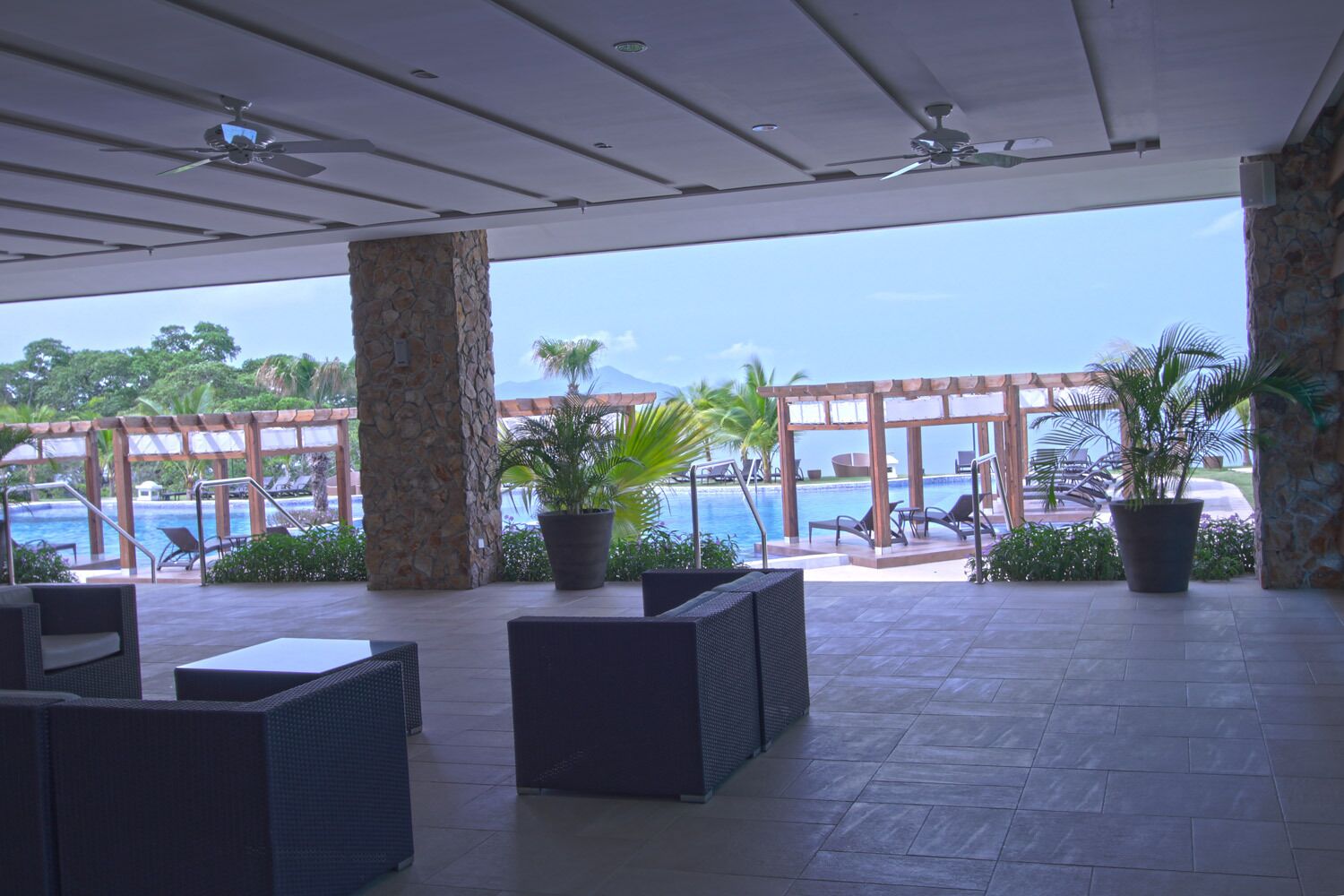 A covered outdoor area with lounge chairs, potted plants, and a view of a pool and ocean.