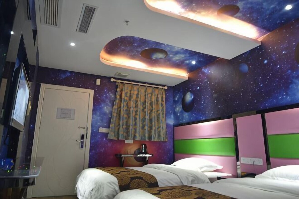 A room with a galaxy-themed wall, a bed with patterned bedding, a bedside table with a lamp, and a television on the left.