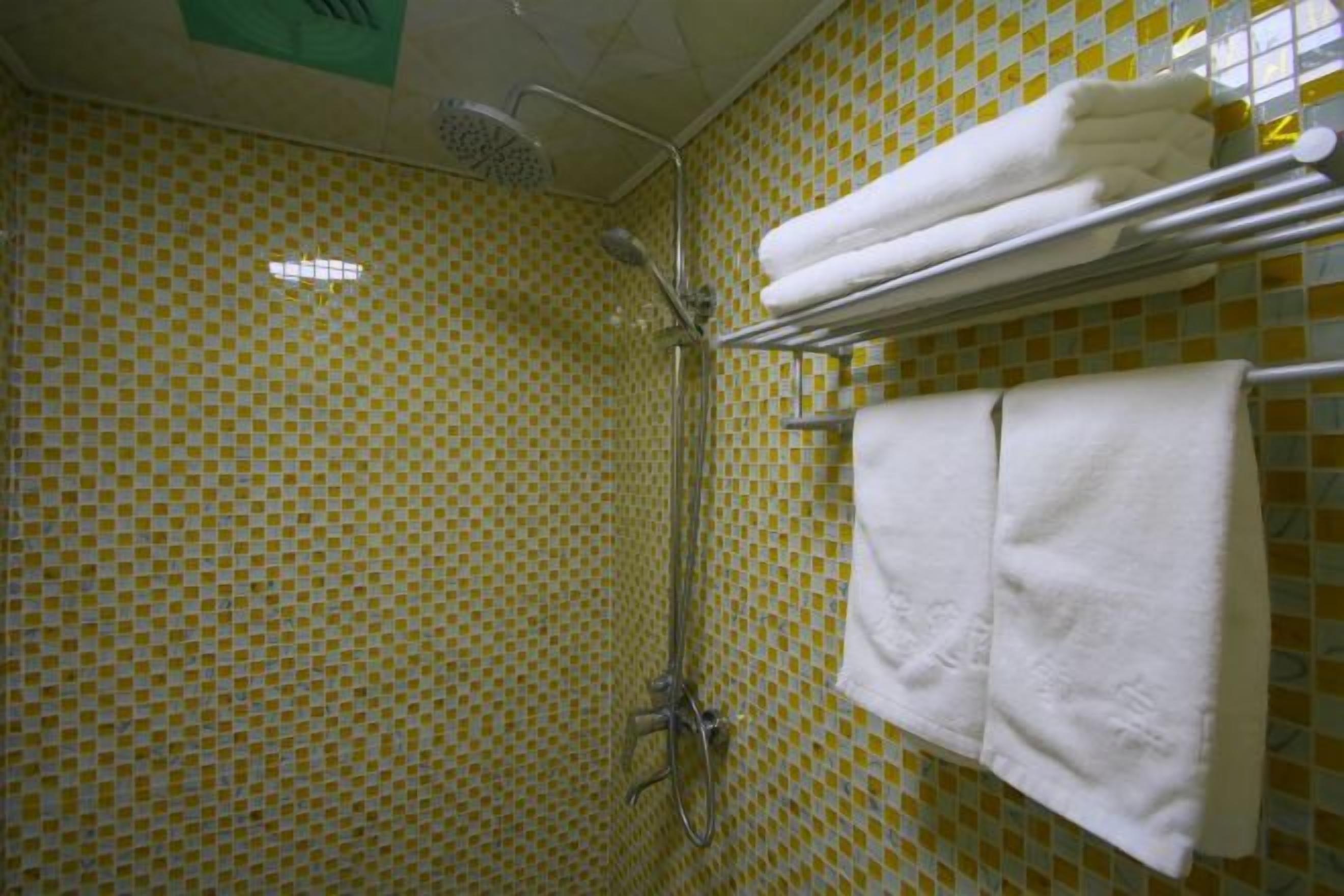 Une salle de bain avec un mur recouvert de carreaux jaunes et blancs formant un motif à carreaux, une douche équipée d’un pommeau de douche à main et un porte-serviettes avec des serviettes pliées.
